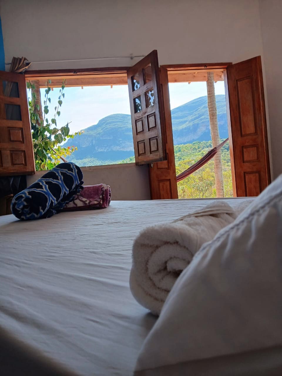 A spacious bedroom features open wooden windows framing a scenic mountain view. Rolled towels are arranged neatly on the bed, complementing the serene atmosphere created by touches of greenery visible outside.