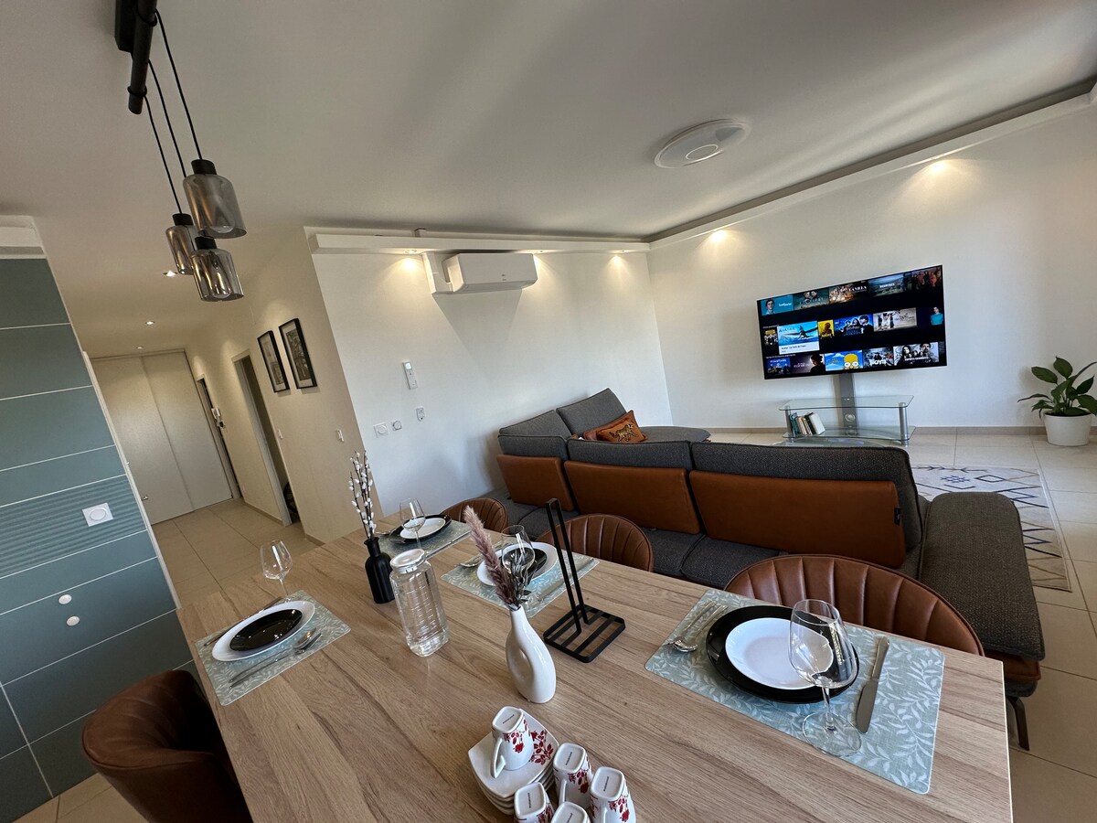 An open-plan living area showcases a modern gray sofa facing a wall-mounted television. A dining table with elegant place settings for four is visible, along with decorative elements including a plant and soft lighting throughout the space.