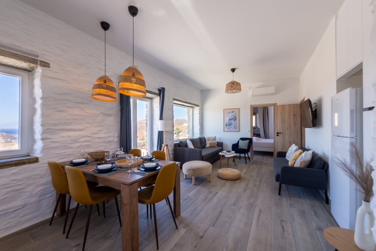 A bright living space features a wooden dining table set for six, surrounded by upholstered chairs. Two pendant lights hang above the table. A cozy seating area with a couch and armchair is positioned to the right, with windows providing views of the outdoors.