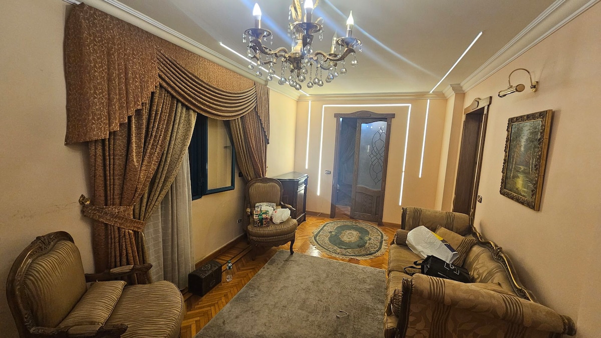 The living area features an elegant chandelier above a plush carpet, with two armchairs positioned around a small table. Soft drapes frame the windows, and a decorative rug is placed on the wooden floor. A door leads to the outside, enhancing light within the space.