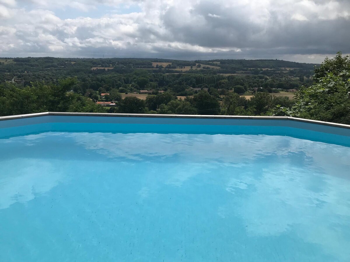 A spacious solar-heated pool reflects the sky, surrounded by lush greenery. The pool's edge seems to blend with the horizon, offering panoramic views of rolling hills and valleys in the distance under a cloudy sky.