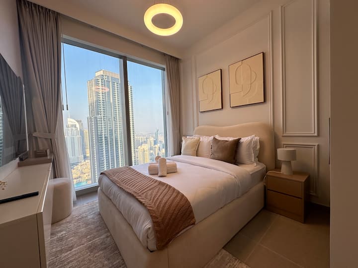 El dormitorio tiene grandes ventanales con impresionantes vistas a la ciudad. Cuenta con una cama acogedora con sábanas blancas y un tiro beige, almohadas decorativas, mesitas de noche con lámparas y arte abstracto en las paredes, creando un ambiente moderno y sereno.
