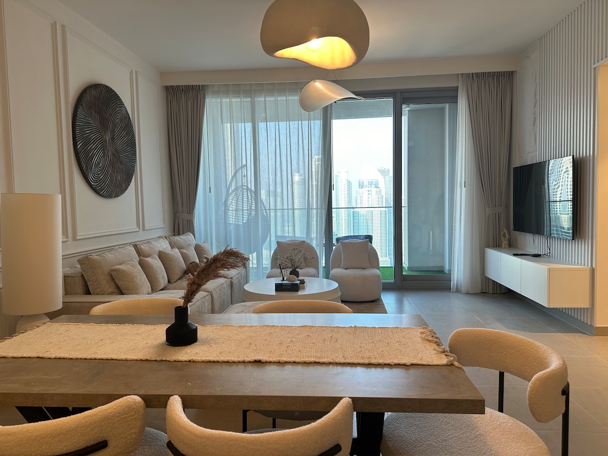 החדר כולל עיצוב מודרני ומאוורר עם לוח צבעים בהיר. ספה נוחה וכיסאות מסוגננים מקיפים שולחן קפה עגול. שולחן אוכל עם כיסאות רכים יושב בקרבת מקום, עם תאורה אלגנטית וחלונות גדולים המציעים נוף לעיר.