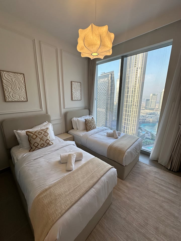 La habitación cuenta con dos camas cuidadosamente hechas con sábanas blancas suaves y almohadas decorativas. Una elegante luz colgante cuelga arriba, iluminando el espacio. Los grandes ventanales ofrecen una vista impresionante de los rascacielos, creando un ambiente luminoso y moderno.