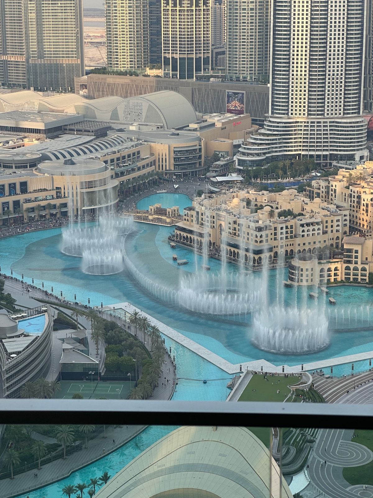 A stunning aerial view captures the Dubai Fountain's elegant water display, featuring multiple streams creating a dynamic pattern against the backdrop of the vibrant cityscape. The surrounding architecture and lush green areas enhance the captivating scene of Downtown Dubai.