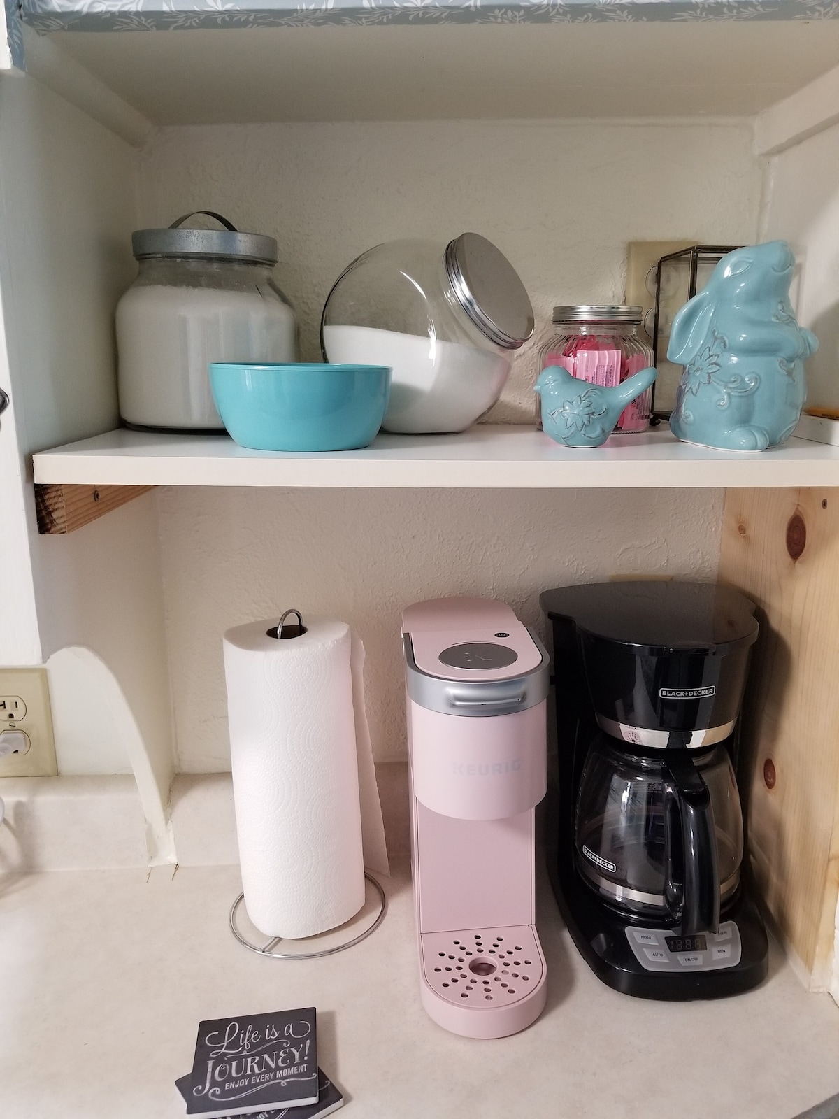 A compact kitchen counter is displayed with various items, including a pink coffee maker and a black coffee machine. A light blue bowl and jar are positioned next to a decorative rabbit, accompanied by a ceramic container and paper towels on a holder.