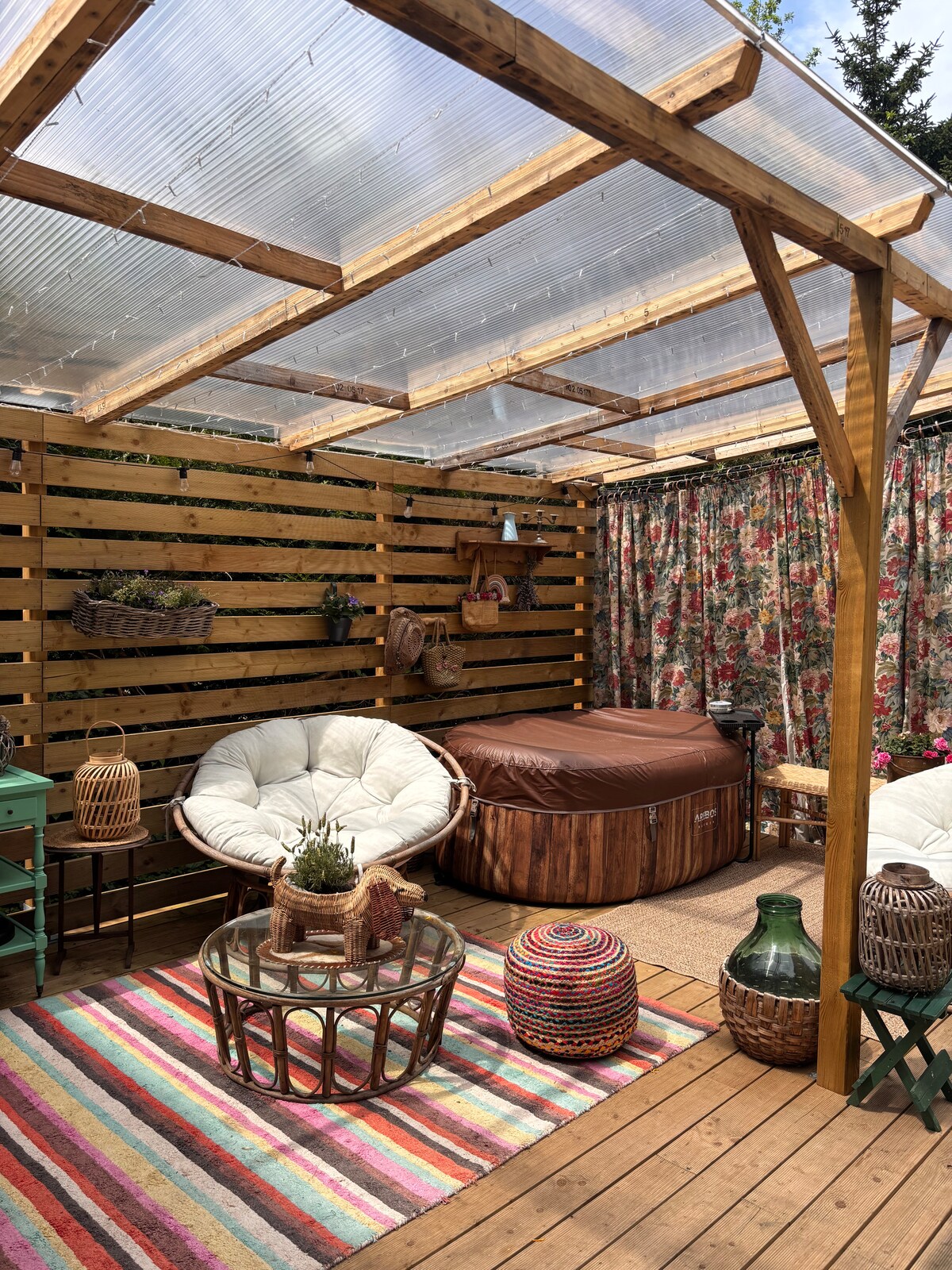 A sheltered outdoor space features a round wooden hot tub and two comfortable chairs styled with soft cushions. A vibrant rug spans the area, complemented by a glass table and decorative elements. Colorful curtains and greenery enhance the natural setting.
