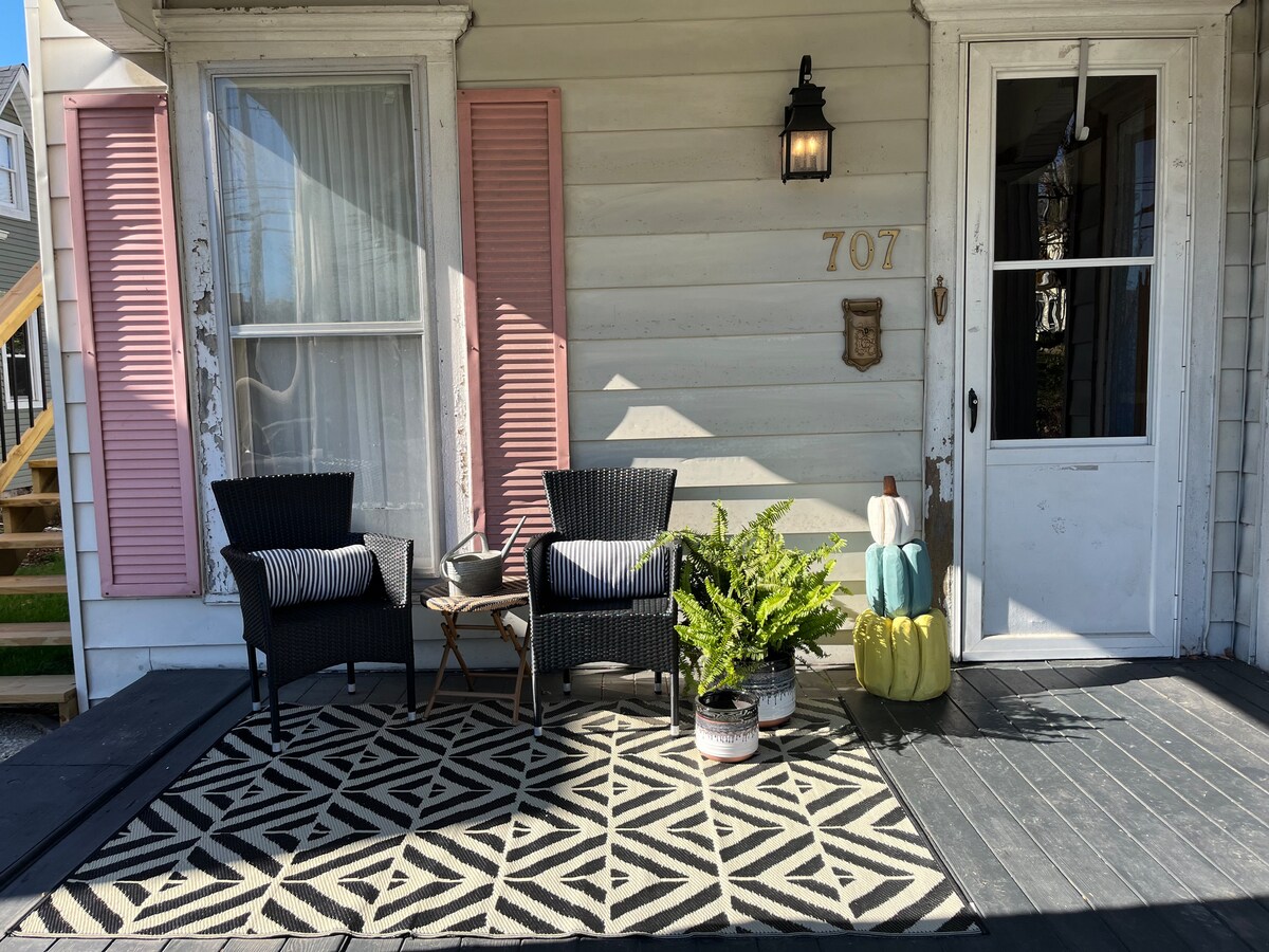 A welcoming front porch features two black wicker chairs with striped cushions, positioned around a small side table. A patterned rug lies underfoot, and potted plants add greenery. The entrance is framed by pink shutters and a light fixture, enhancing the charming exterior.