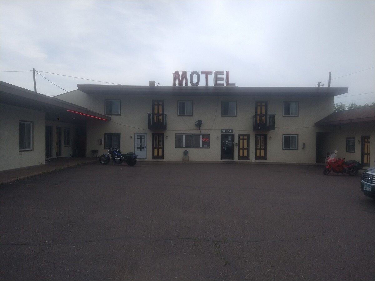 A modest motel building with a prominent red 'MOTEL' sign is positioned at the top. The two-story structure showcases several balconies with yellow doors. A paved parking area is visible in front, accommodating motorcycles and vehicles.