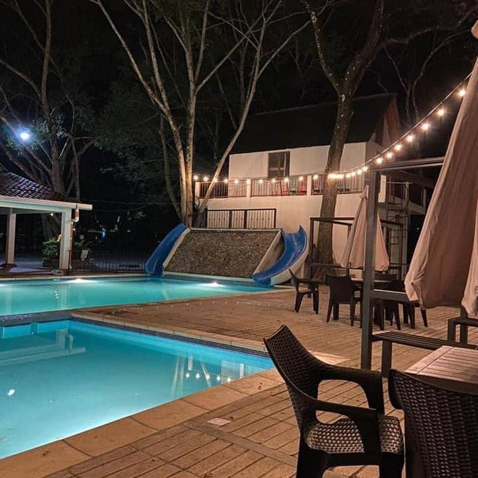 A nighttime view features a serene swimming pool with blue accents, supplemented by a water slide nearby. String lights provide gentle illumination over outdoor seating arrangements comprised of tables and chairs. The surrounding area is framed by trees, creating a calm atmosphere.