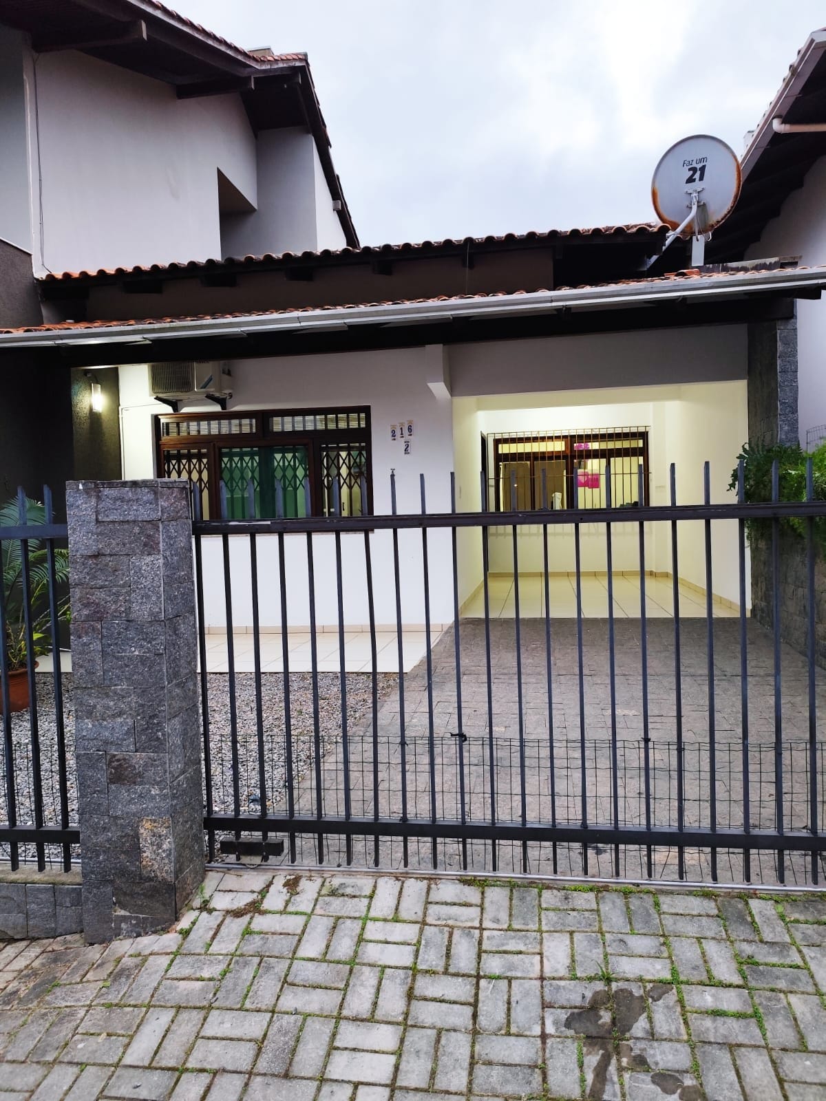 The property features a gated entrance with a stone fence, leading to a gravel pathway. The facade is adorned with green windows and a spacious garage area. Light grey walls are complemented by a tiled roof, creating a welcoming appearance.