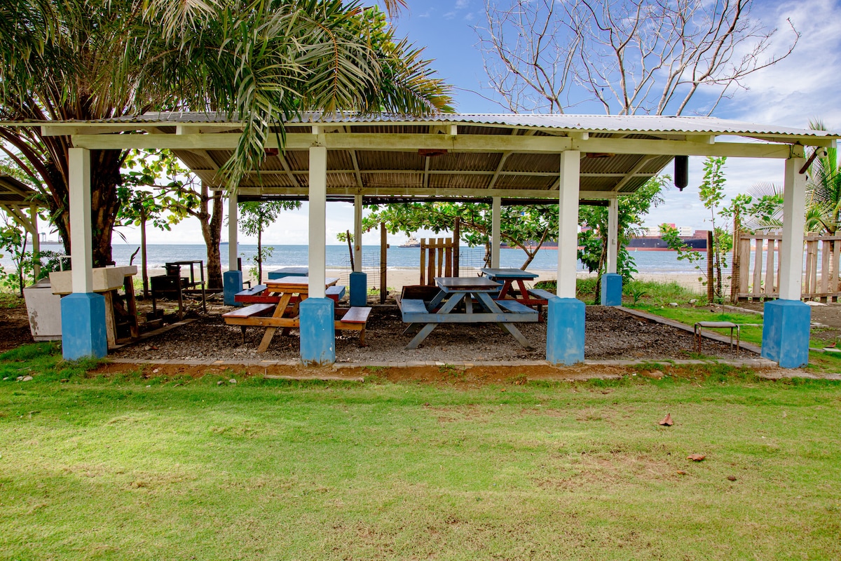 An open pavilion features picnic tables with benches, surrounded by green grass and palm trees. The structure's roof provides shade, while the nearby ocean can be seen in the background, adding a serene coastal ambiance to the outdoor dining area.