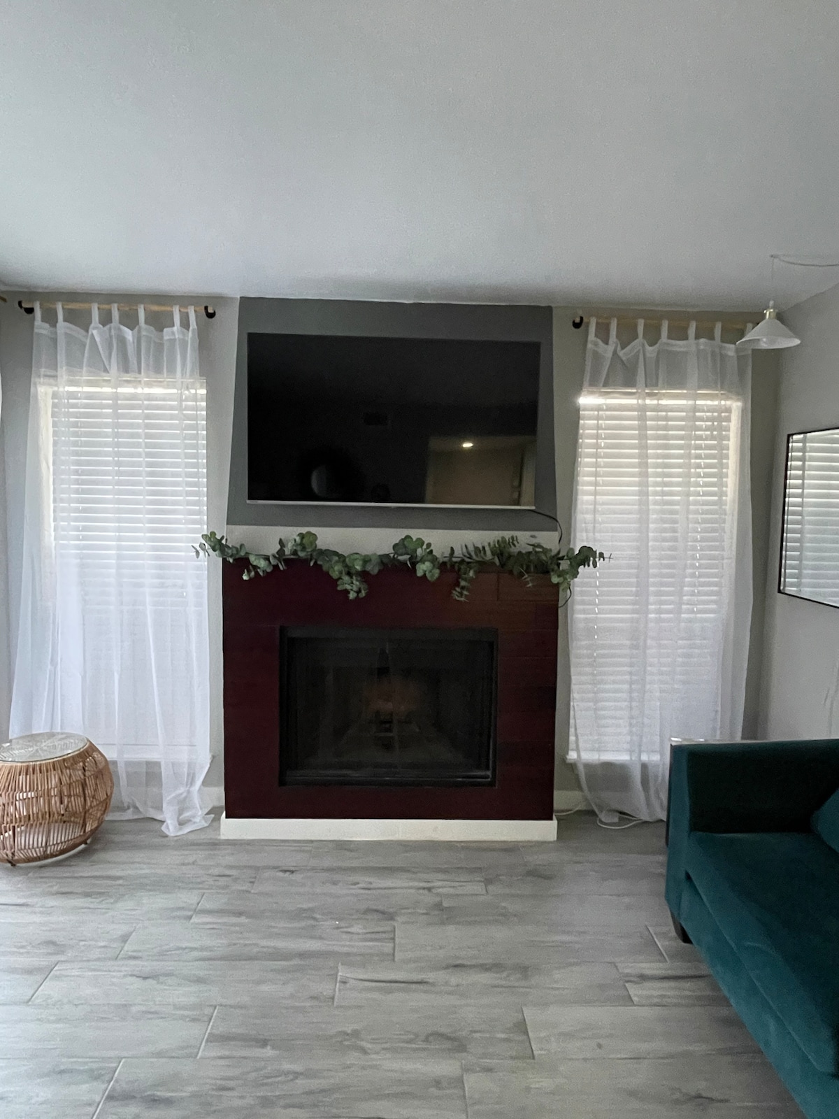 A modern living area features a sleek fireplace with a dark wood mantel, adorned with greenery. Two large windows with sheer white curtains allow natural light, and a comfortable teal sofa provides a seating area. A subtle decorative mirror is positioned nearby.