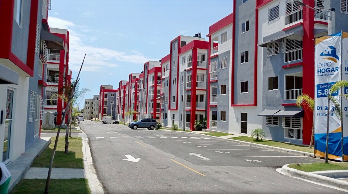 A view of a modern residential complex is displayed, featuring multiple multi-story buildings painted in red and gray. A well-maintained asphalt road runs through the center, flanked by neatly trimmed grass and palm trees, suggesting a welcoming environment.