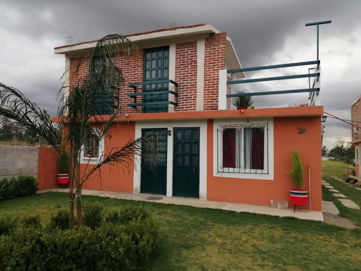 A two-story house is depicted, featuring a combination of orange and brick exterior. A green balcony with railings is visible on the upper floor, while a well-maintained lawn with shrubs surrounds the base. Large windows and a double door entrance enhance the inviting façade.