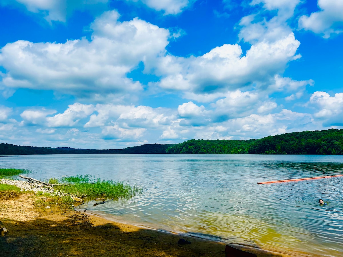 The image captures a serene lakeside view under a bright blue sky, featuring soft clouds above calm water. Lush greenery frames the lake, with sandy areas visible at the shoreline. The tranquil atmosphere reflects the natural beauty of the surrounding landscape.
