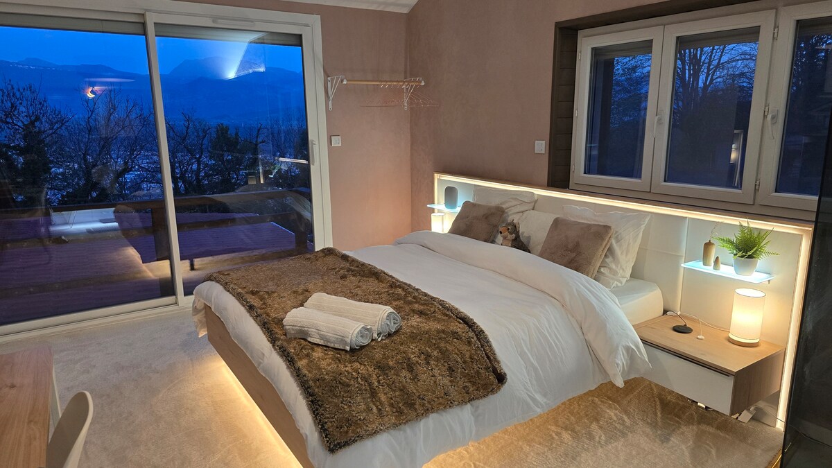 A cozy bedroom features a spacious bed, softly illuminated by built-in lighting. Large windows provide views of the outdoor landscape, while a warm rug adds a touch of comfort. A bedside table holds a small plant, and a set of towels is neatly arranged on the bed.