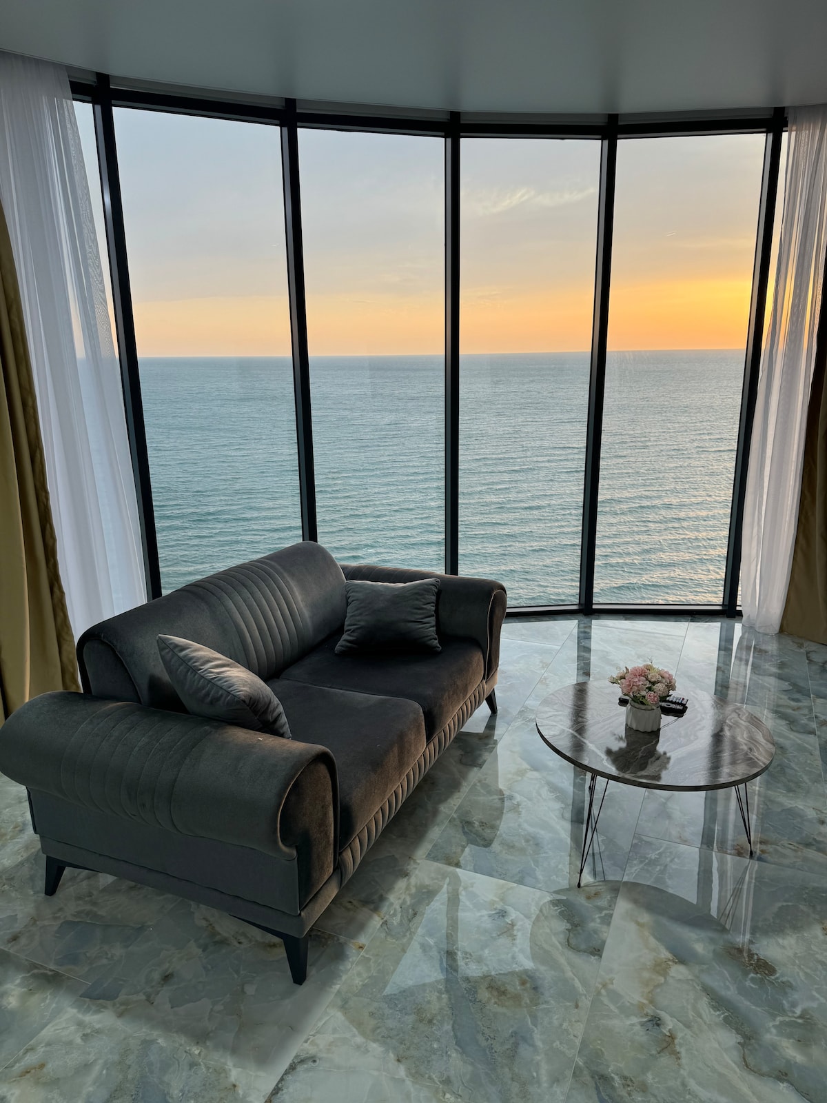 A comfortable sofa is positioned beside expansive floor-to-ceiling windows, offering a panoramic view of the sea at sunset. A small glass table with a vase of flowers is nearby, enhancing the serene ambiance of the space.
