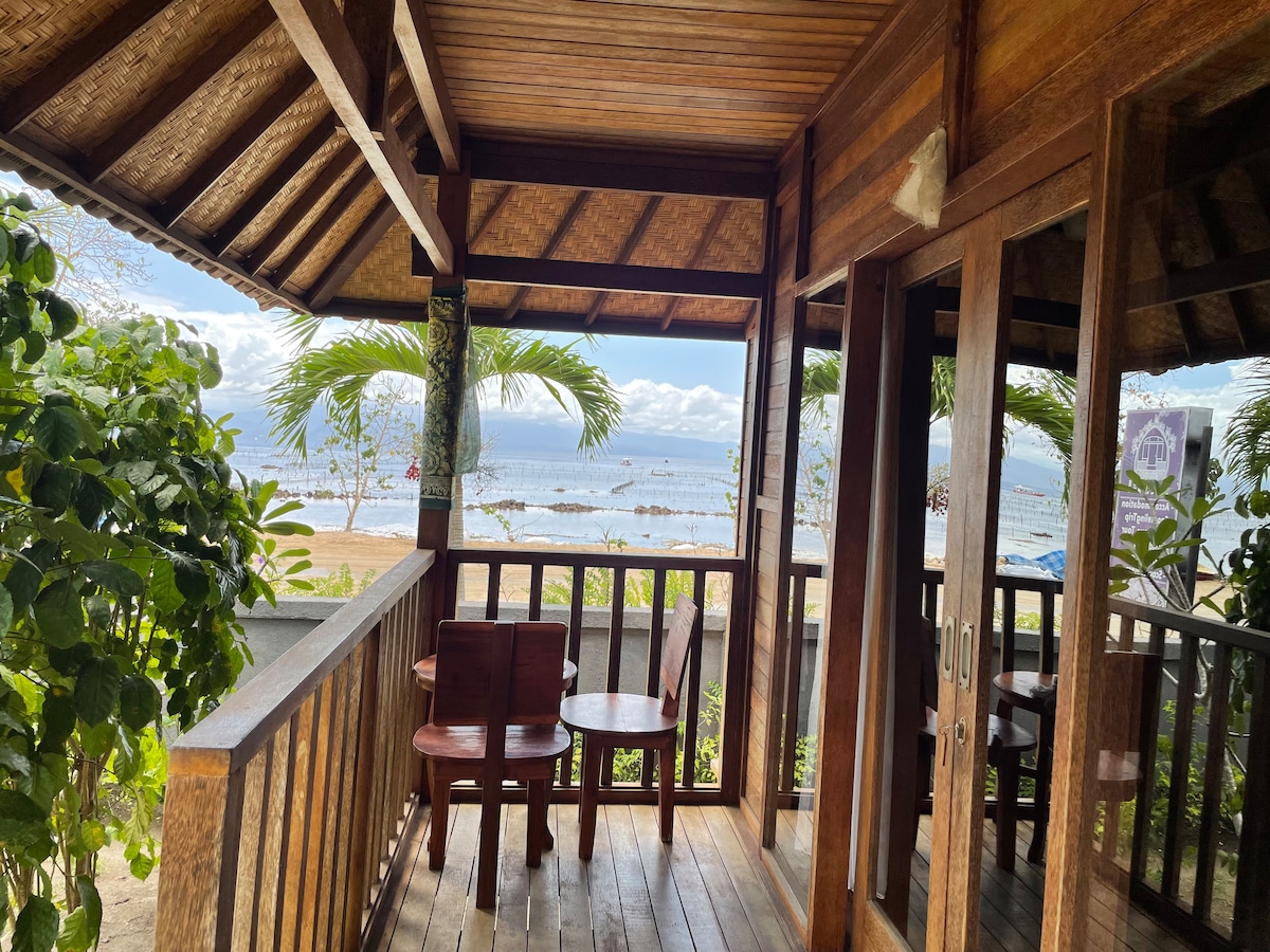 A wooden balcony offers views of the beach, framed by lush greenery. Two chairs and a small table are positioned invitingly on the wooden deck, while sunlight reflects off the water. The thatched roof adds a traditional touch to the outdoor space.