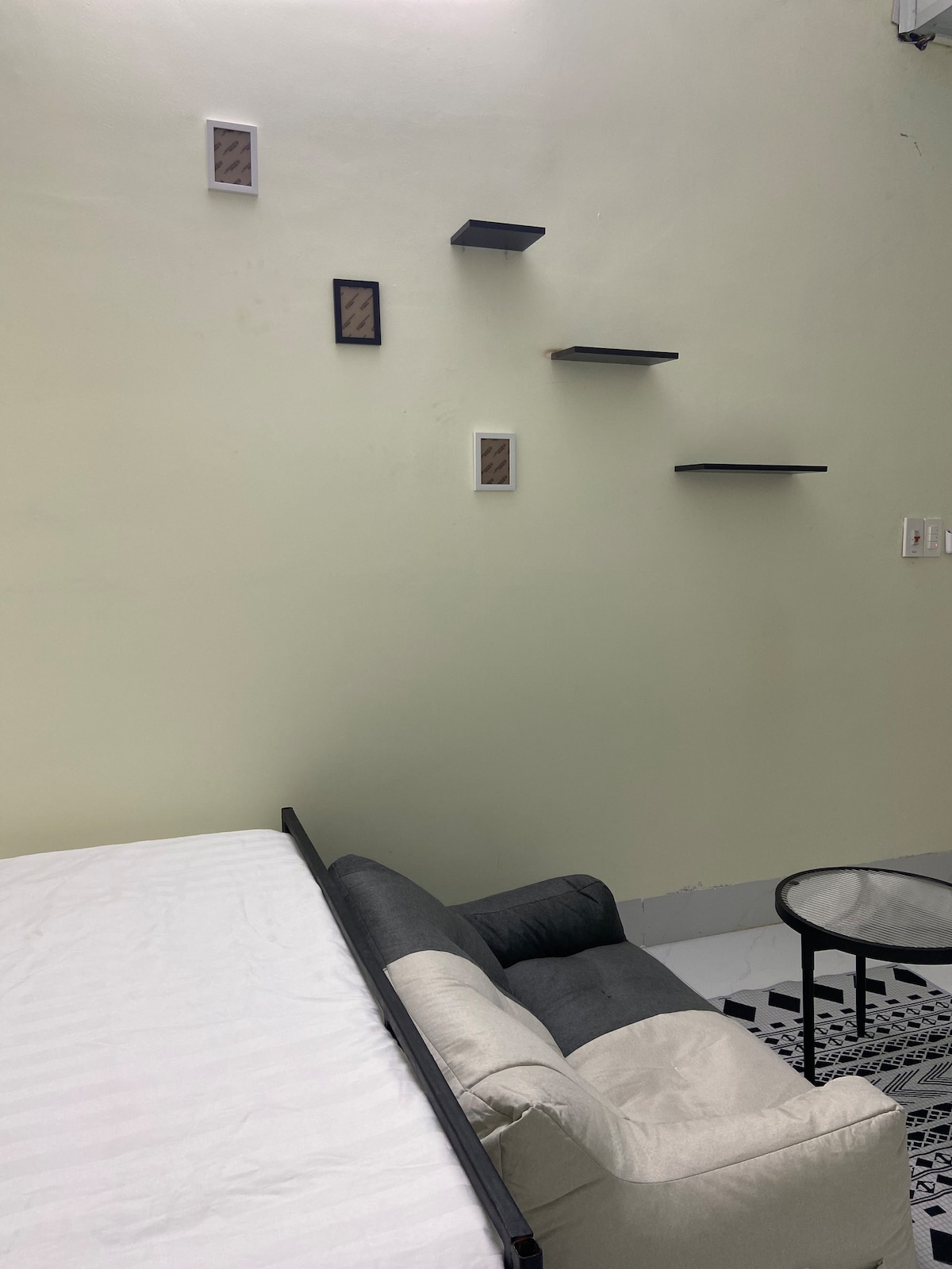 A simple sleeping area is complemented by a low, neutral-toned couch and a small table. Decorative wall shelves in varying sizes are mounted above, enhancing the space's vertical appeal. A light-colored bed occupies the foreground, creating a relaxed environment.