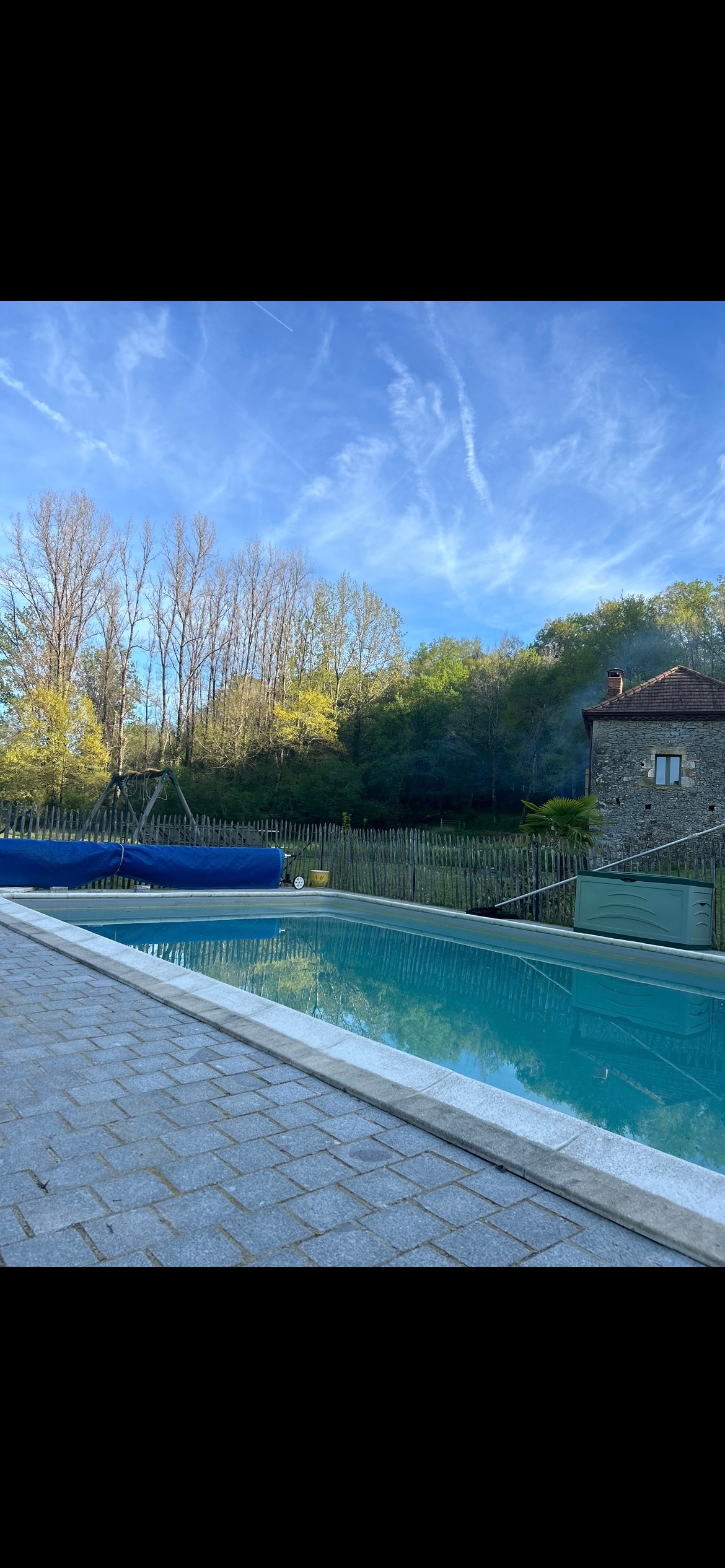 A tranquil swimming pool is set against a backdrop of lush trees and blue skies. The pool's clear water reflects the surrounding greenery. A paved area surrounds the pool, offering space for relaxation. A rustic stone building is visible in the background.