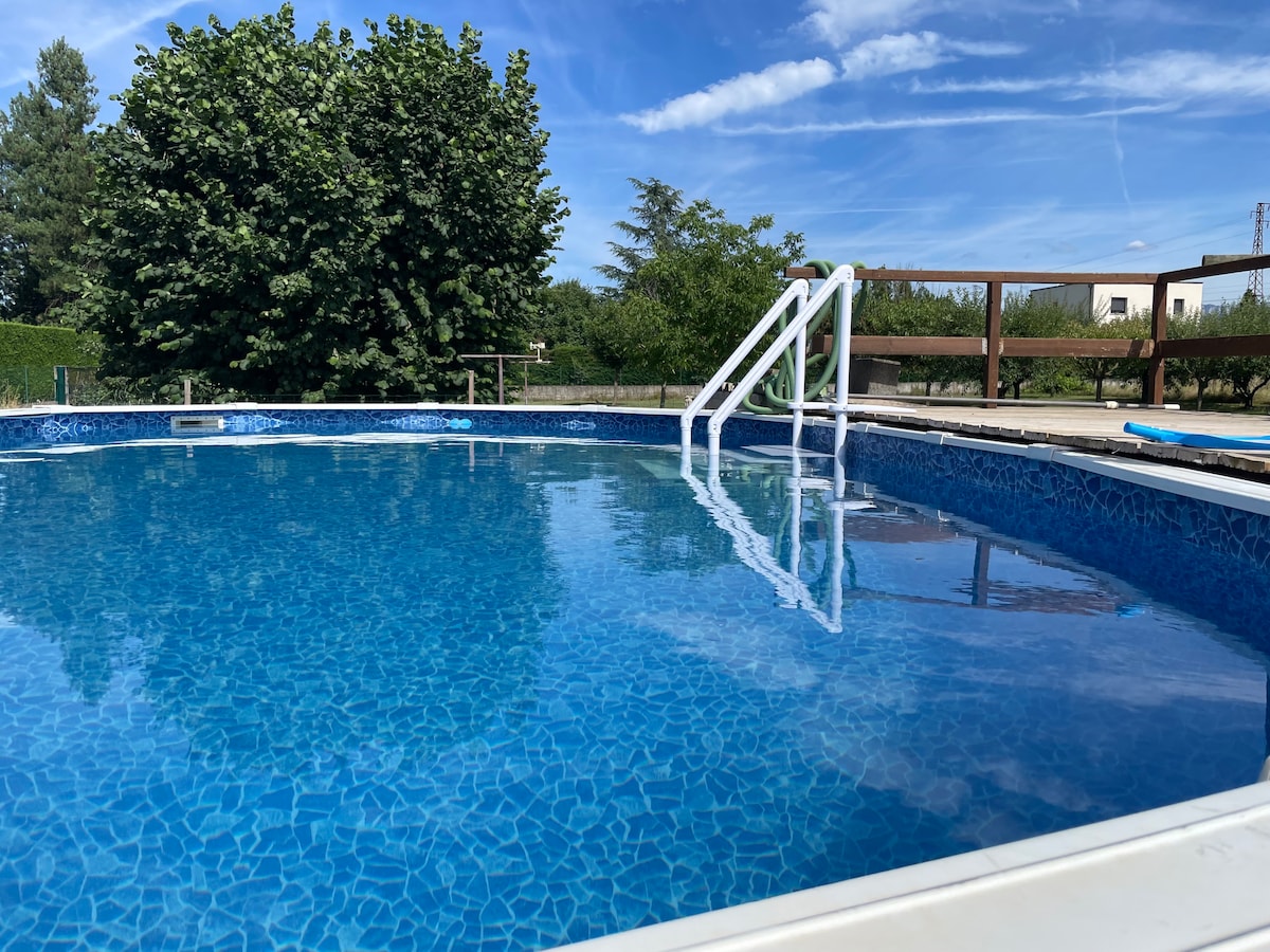 An above-ground pool features clear blue water, reflecting the sky. A set of steps leads into the water, surrounded by a wooden deck. Lush greenery lines the background, creating a serene outdoor environment.