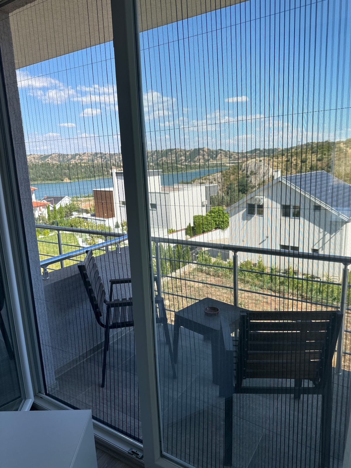 A balcony view is showcased, featuring a small table and two chairs with a lake and surrounding hills visible in the background. The mesh screen filters light, creating a soft atmosphere while maintaining a connection with the outdoor scenery.