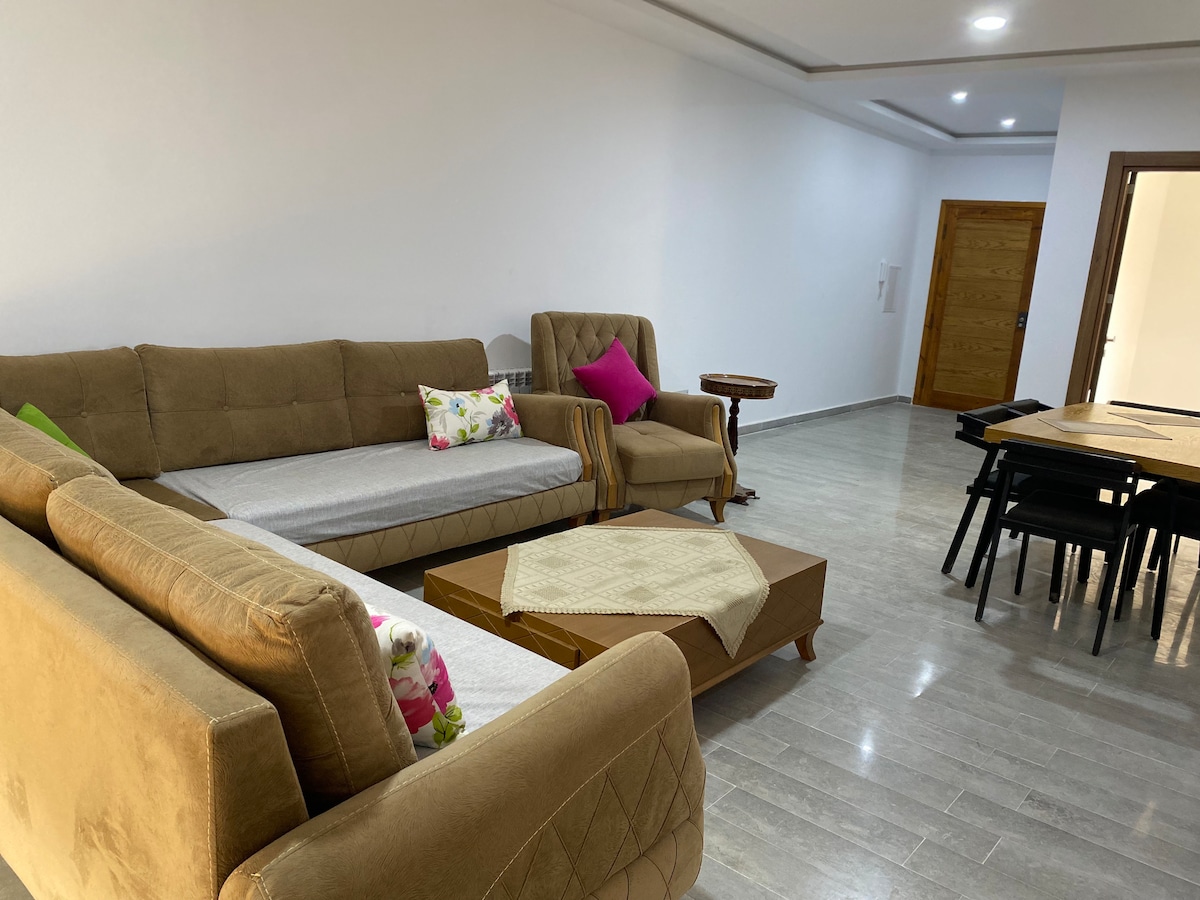 A spacious living area features a combination of comfortable seating, including two sofas and a chair adorned with colorful cushions. A coffee table sits at the center, while a dark dining table can be seen in the background near an inviting wooden door.