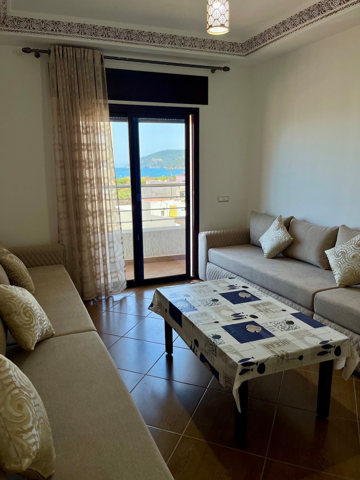 A light-filled living area features two comfortable sofas arranged around a patterned coffee table. Floor-to-ceiling glass doors provide access to a balcony with sea views. Decorative pillows add subtle elegance, while intricate ceiling details enhance the traditional Moroccan design.