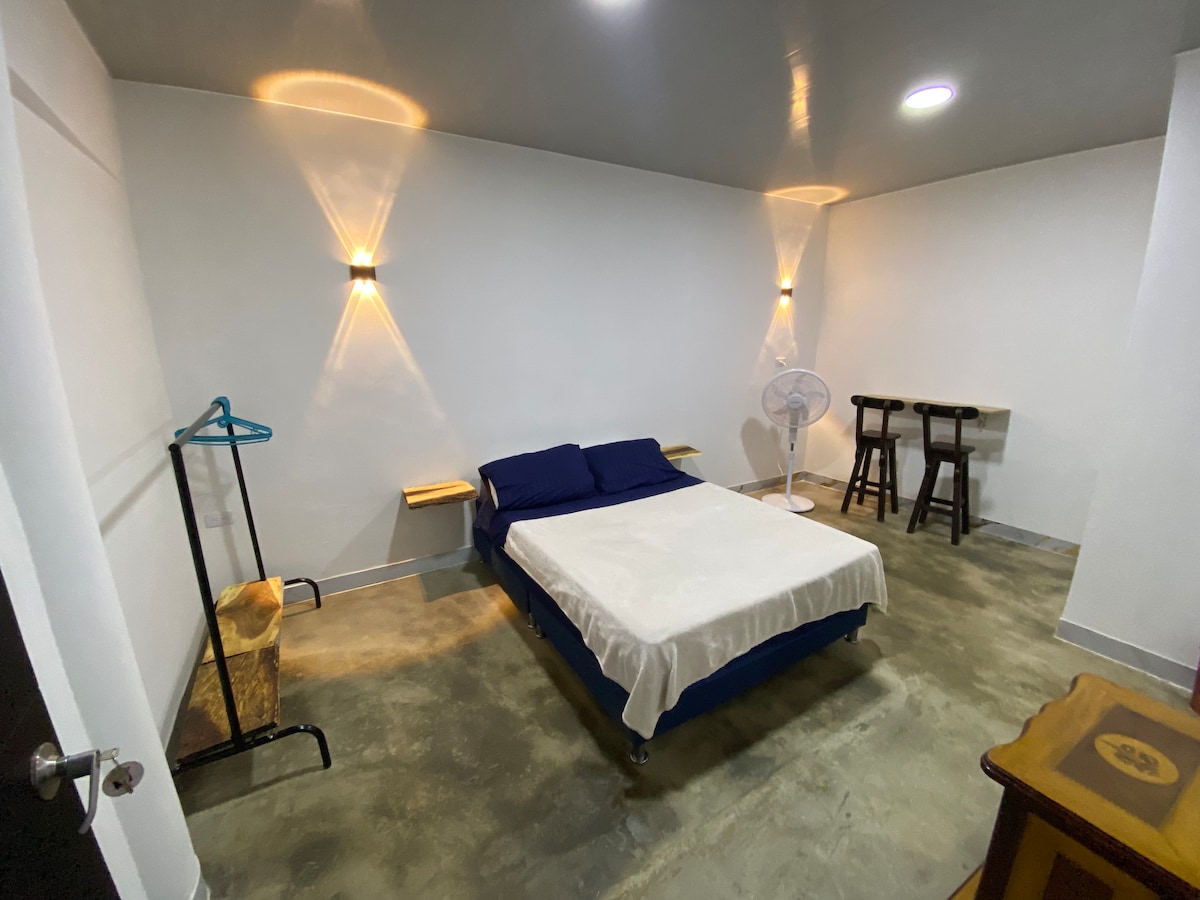 A spacious bedroom is featured with a queen-sized bed adorned with a light-colored blanket. Two minimalist shelves are mounted on the walls, and a small table with two stools is present. A fan provides airflow, while a clothes rack adds utility to the space.