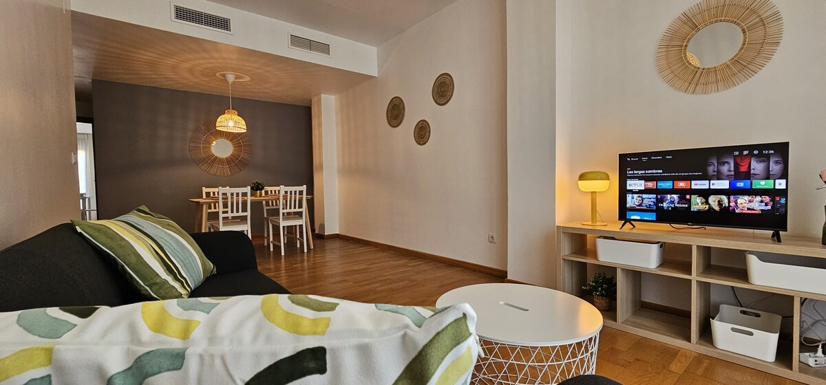 A living area with a modern design features a comfortable sofa adorned with patterned cushions. A light-colored coffee table sits in front, and a flat-screen television stands on a nearby console. A dining table with white chairs is visible in the background.