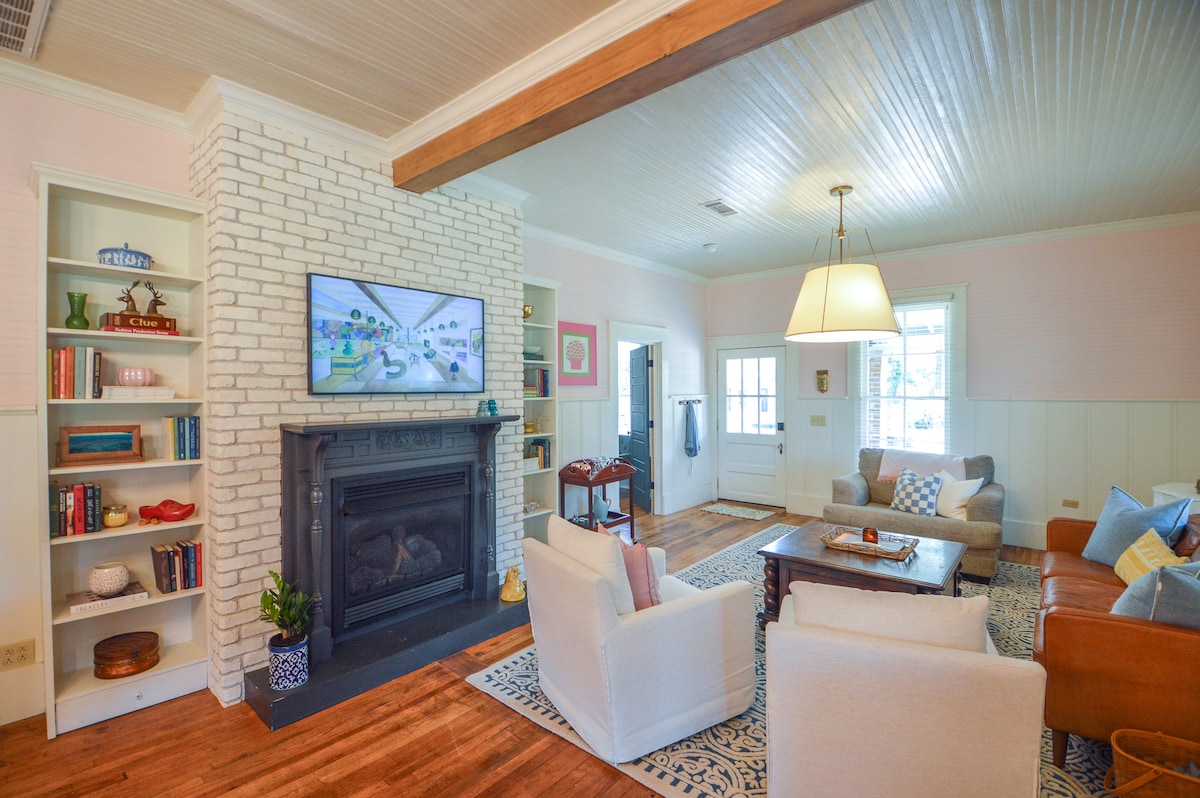 A spacious living area is highlighted by a white brick fireplace and a mounted television. Comfortable seating options include a leather sofa and plush armchairs. Natural light fills the room through large windows, complemented by built-in shelves filled with books and decorative items.