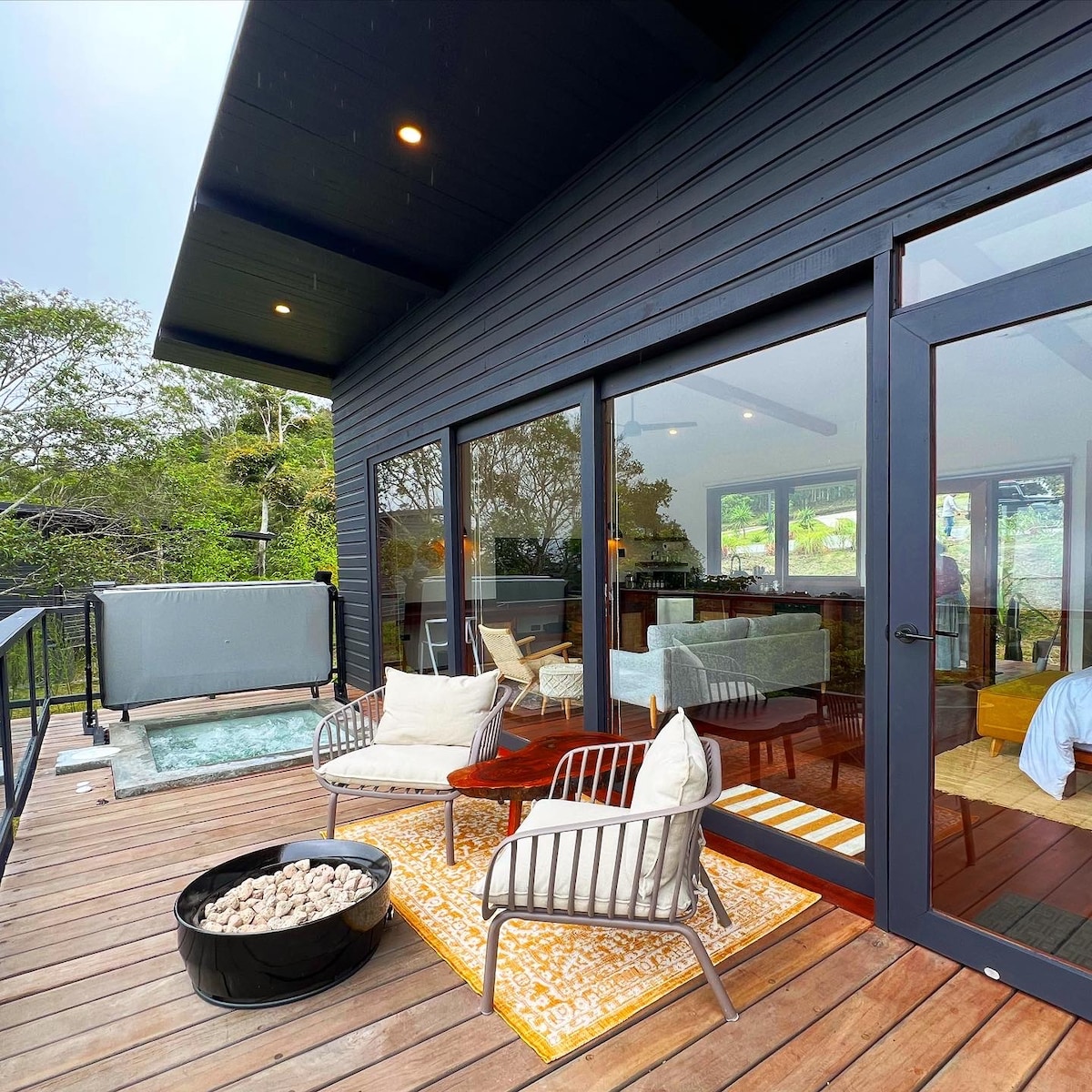 A private deck features two modern chairs with a small table between them, positioned on a textured area rug. A custom-built jacuzzi is visible in the background, surrounded by a wooden railing. Natural light floods the indoor living area through expansive glass doors.