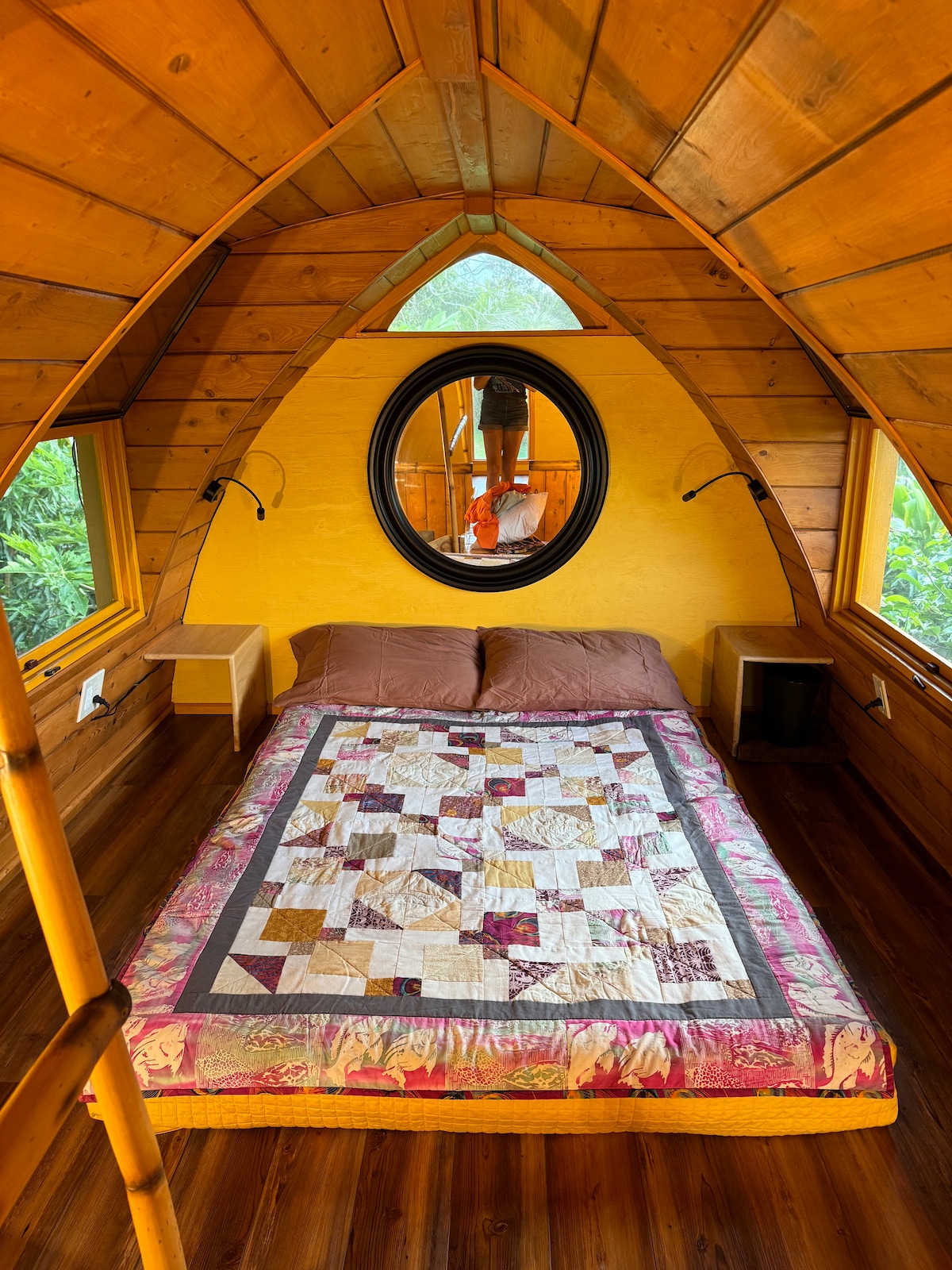 A cozy sleeping area is highlighted by warm wood paneling and an arched ceiling. A comfortable bed is centered beneath a decorative quilt, with two bedside tables flanking either side. A round mirror reflects natural light from a window, enhancing the space.