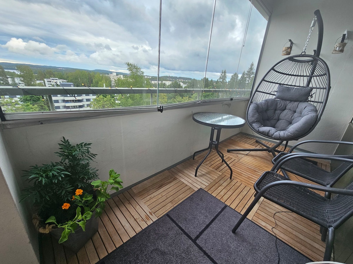 A balcony area is presented with a hanging chair and a small round table. A potted plant with vibrant flowers adds a touch of greenery. Wooden flooring contrasts against a dark rug, enhancing the outdoor space with a relaxed atmosphere.