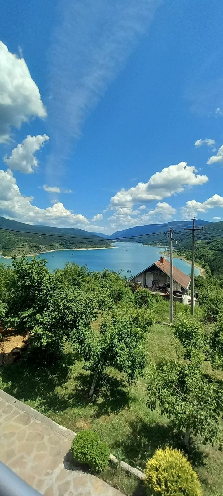 A scenic view captures the tranquil lake surrounded by lush greenery and distant mountains. A quaint house is situated near the water's edge, framed by trees. The sky is filled with scattered clouds, enhancing the peaceful atmosphere of the landscape.