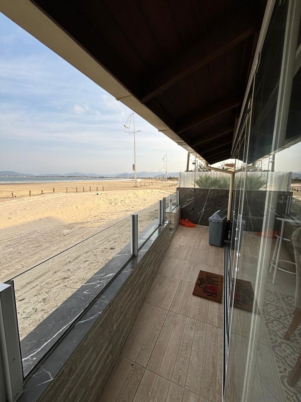 A sandy beachfront view is presented from the patio, framed by a sleek glass railing. The space is bordered by a marble wall, while a small rug adds a touch of comfort. In the distance, beach umbrellas and pathways are visible along the shore.