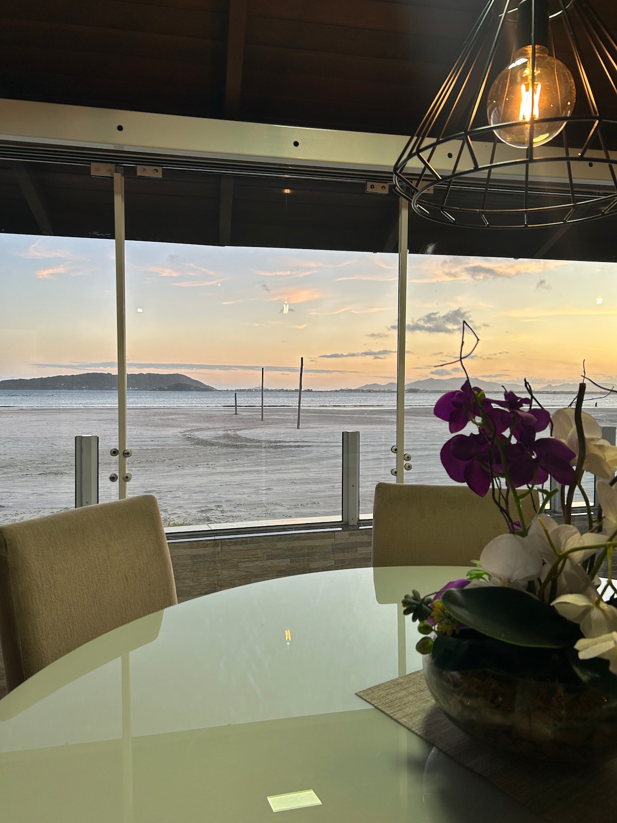 A dining area is featured, showcasing a round table surrounded by light-colored chairs. A floral centerpiece adds a touch of nature. Large glass doors provide an unobstructed view of the beach and sunset, enhancing the connection between indoor dining and the natural scenery.