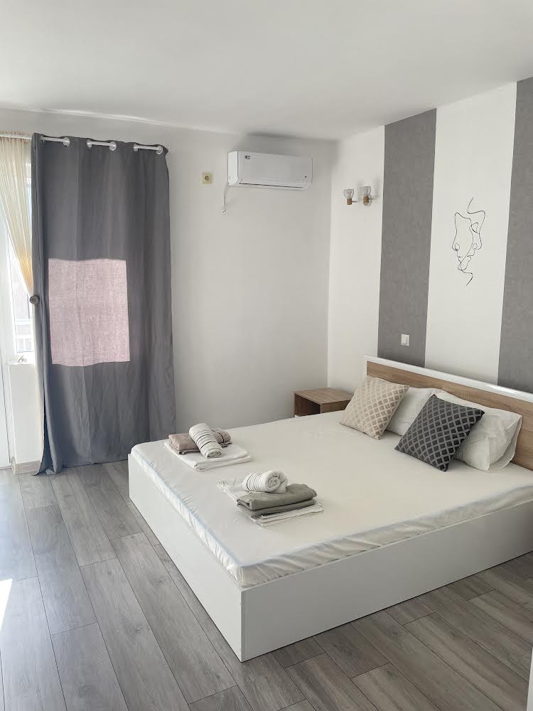 A bright bedroom features a spacious bed with crisp linens and decorative pillows. Light streams through sheer curtains, illuminating the room. A bedside table is positioned next to the bed, and an air conditioning unit is mounted on the wall.