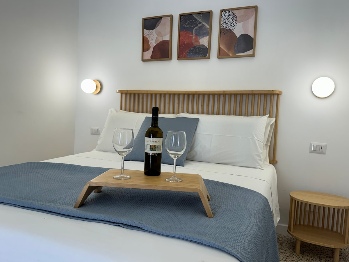 A spacious bedroom features a wooden bed frame with a light-colored mattress and soft bedding. A tray with two glasses and a bottle of wine rests on the bed. Above, three framed artworks add subtle color to the neutral walls, with modern bedside lights flanking the headboard.