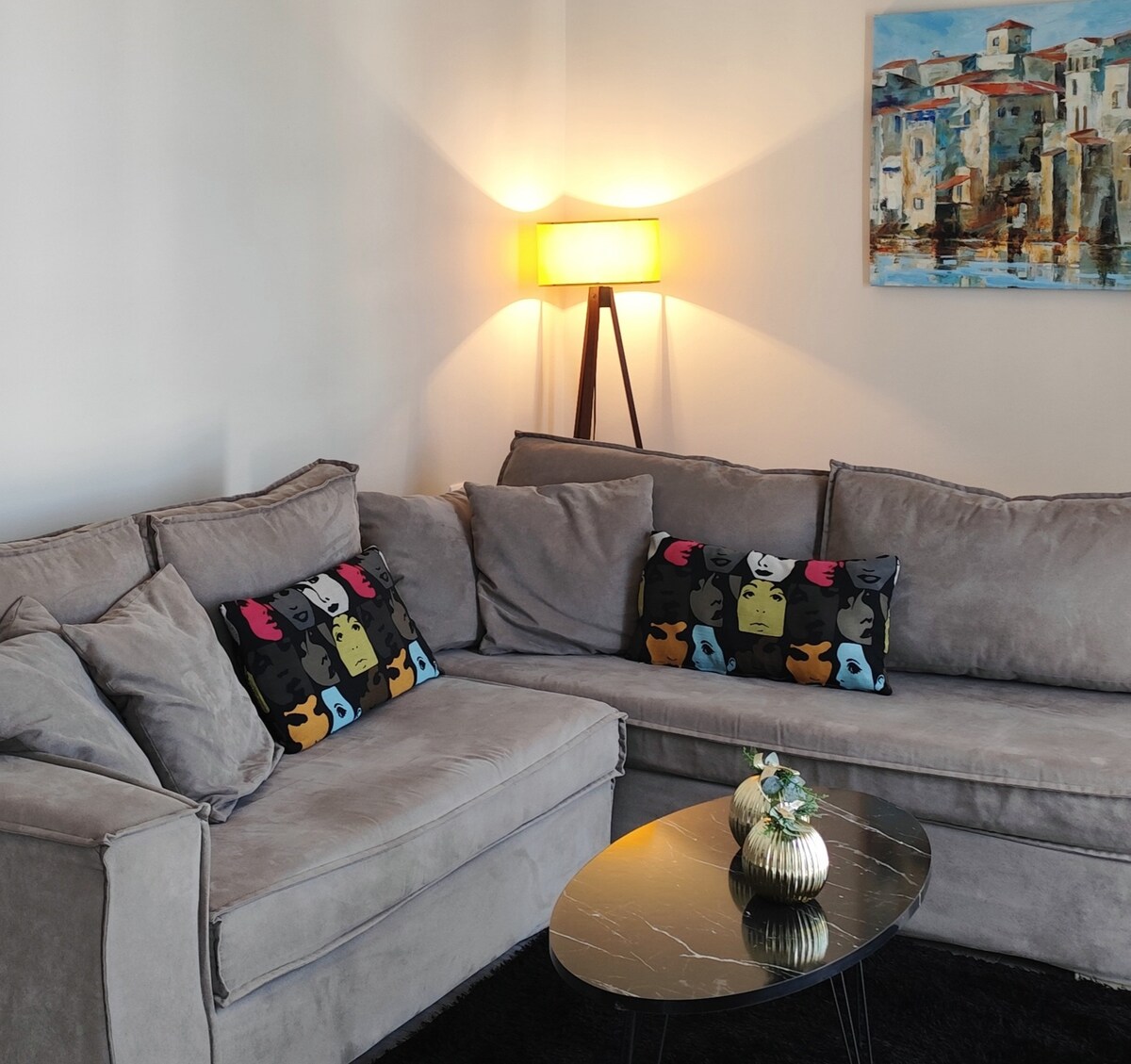 A comfortable corner of the living room features a spacious gray sofa with decorative pillows displaying colorful abstract faces. A small round black coffee table is positioned in front, complemented by a stylish lamp that provides warm lighting. A canvas painting of a coastal scene hangs above.