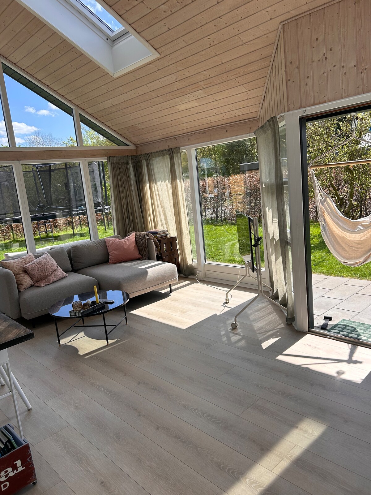 A winter garden is illuminated by natural light, featuring a comfortable sofa and a small coffee table. Large glass doors provide a view of the green garden, while sheer curtains frame the space. A relaxing hammock is visible in the background.