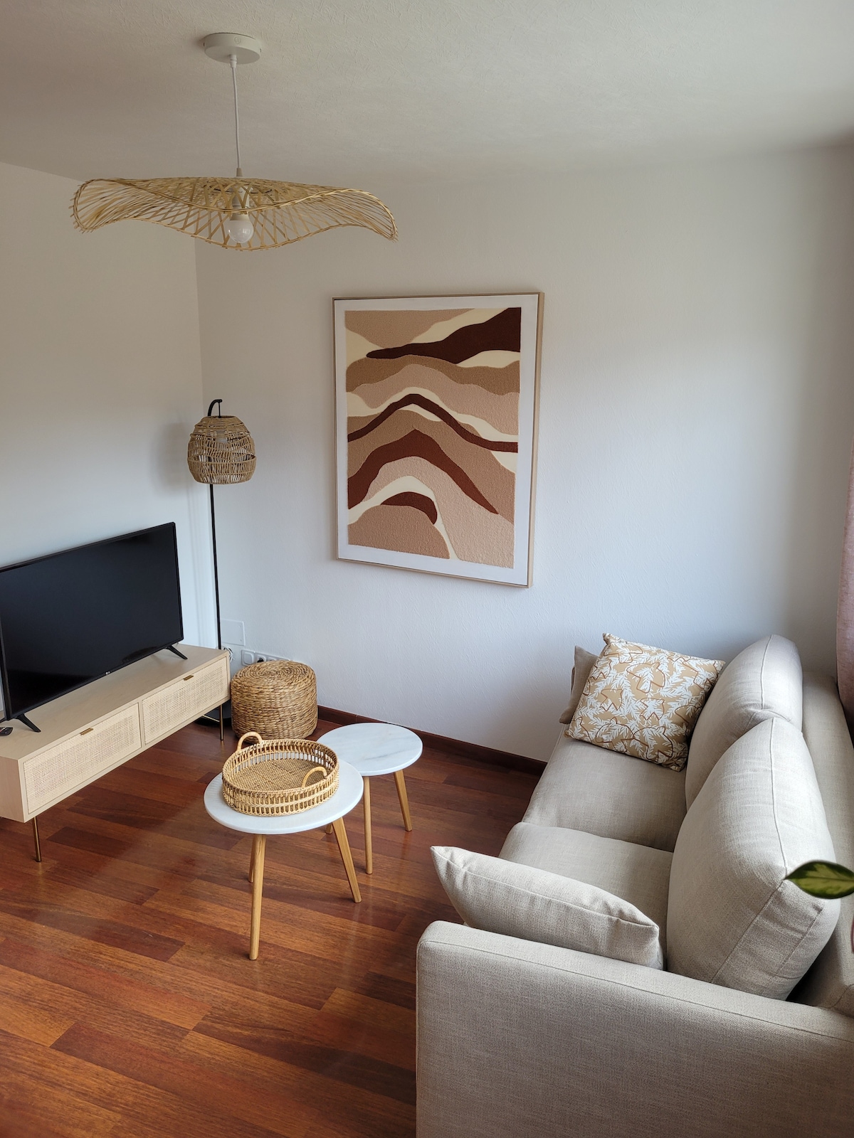 A cozy living area features a light-colored sofa accompanied by two small tables. A TV is placed on a low cabinet, and a decorative wall art piece in warm tones adds visual interest. Natural light enhances the wooden floor, creating a pleasant atmosphere.
