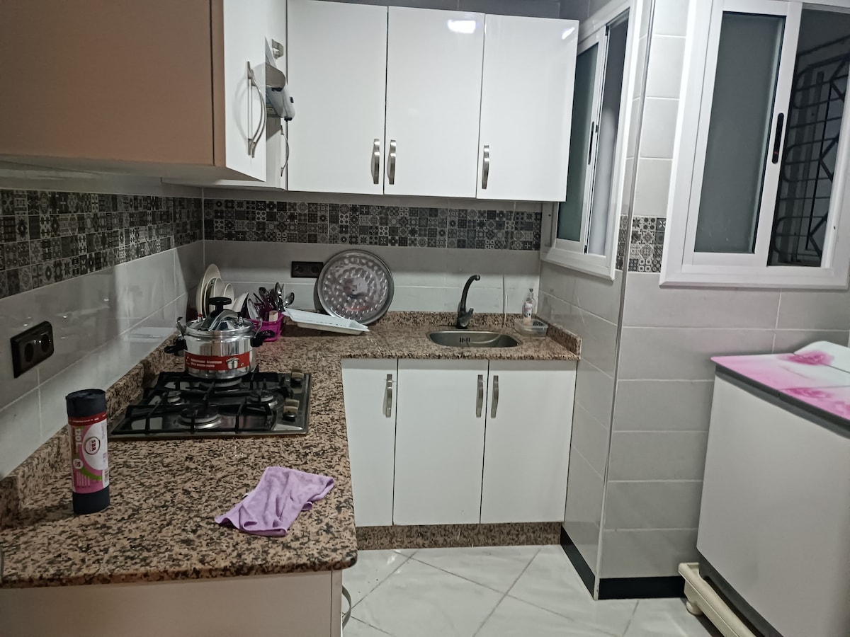A modern kitchen features white cabinetry and a granite countertop. A gas stove is positioned near the sink, which is surrounded by patterned tile backsplash. Natural light enters through two windows, with a drying rack and utensils visible on the counter.