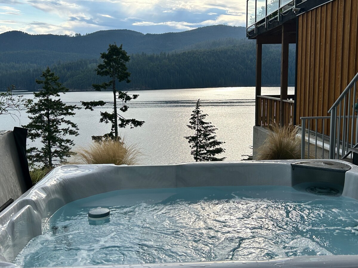 A private hot tub is positioned on the deck, overlooking the serene waters of Sechelt Inlet. The scenic backdrop features lush green trees and distant mountains, creating a peaceful environment for relaxation.