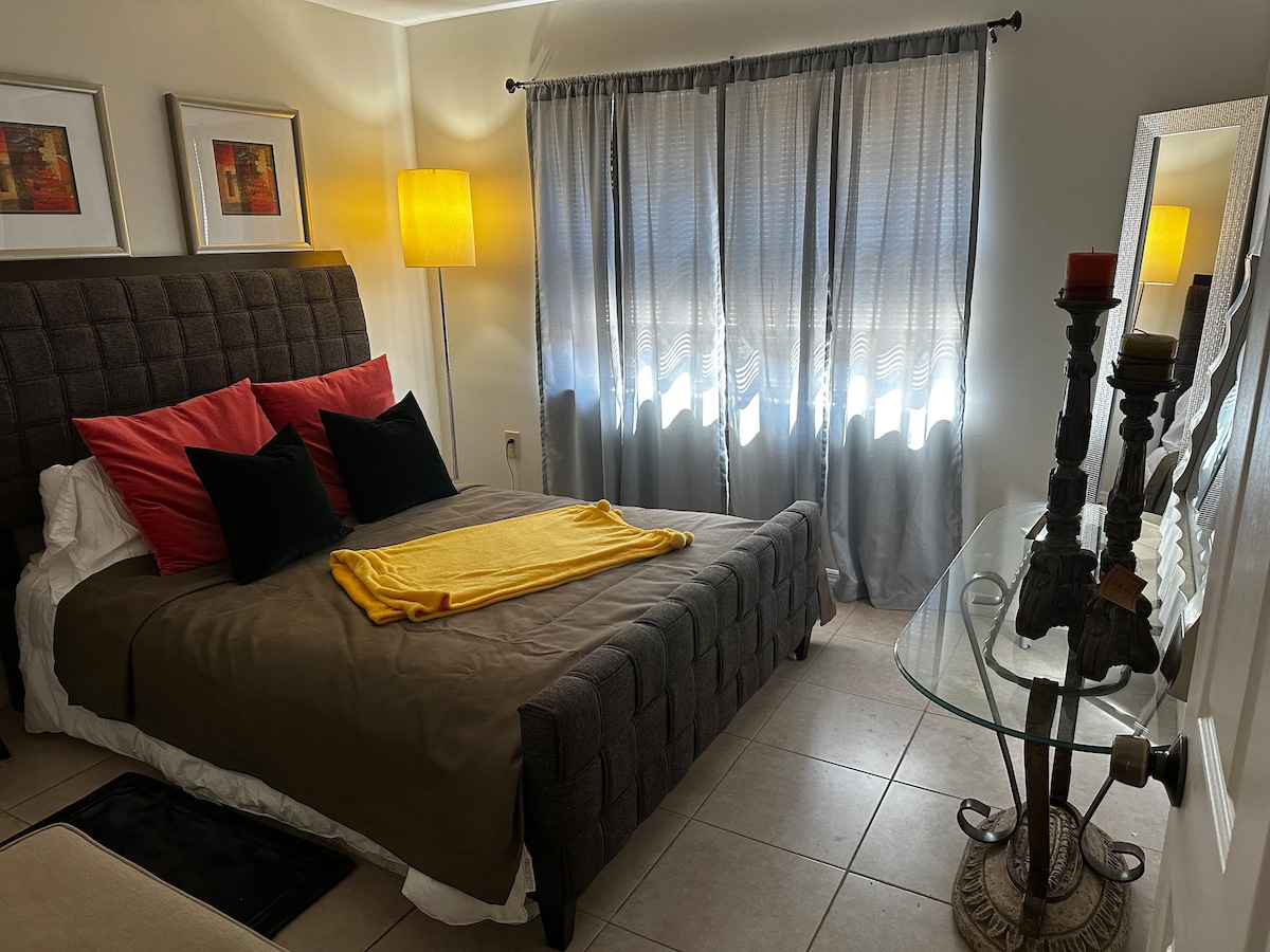A sleeping area features a stylish upholstered bed with red and black accent pillows. A yellow throw blanket rests at the foot of the bed. Light filters through sheer curtains, creating a soft glow. A side table with decorative accents complements the space.