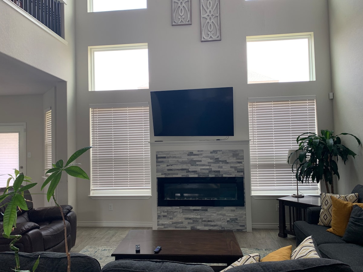 A spacious living area features high ceilings with large windows allowing natural light to fill the room. A mounted television is situated above a modern fireplace, with a coffee table centered on a comfortable seating arrangement. Potted plants add a touch of greenery.