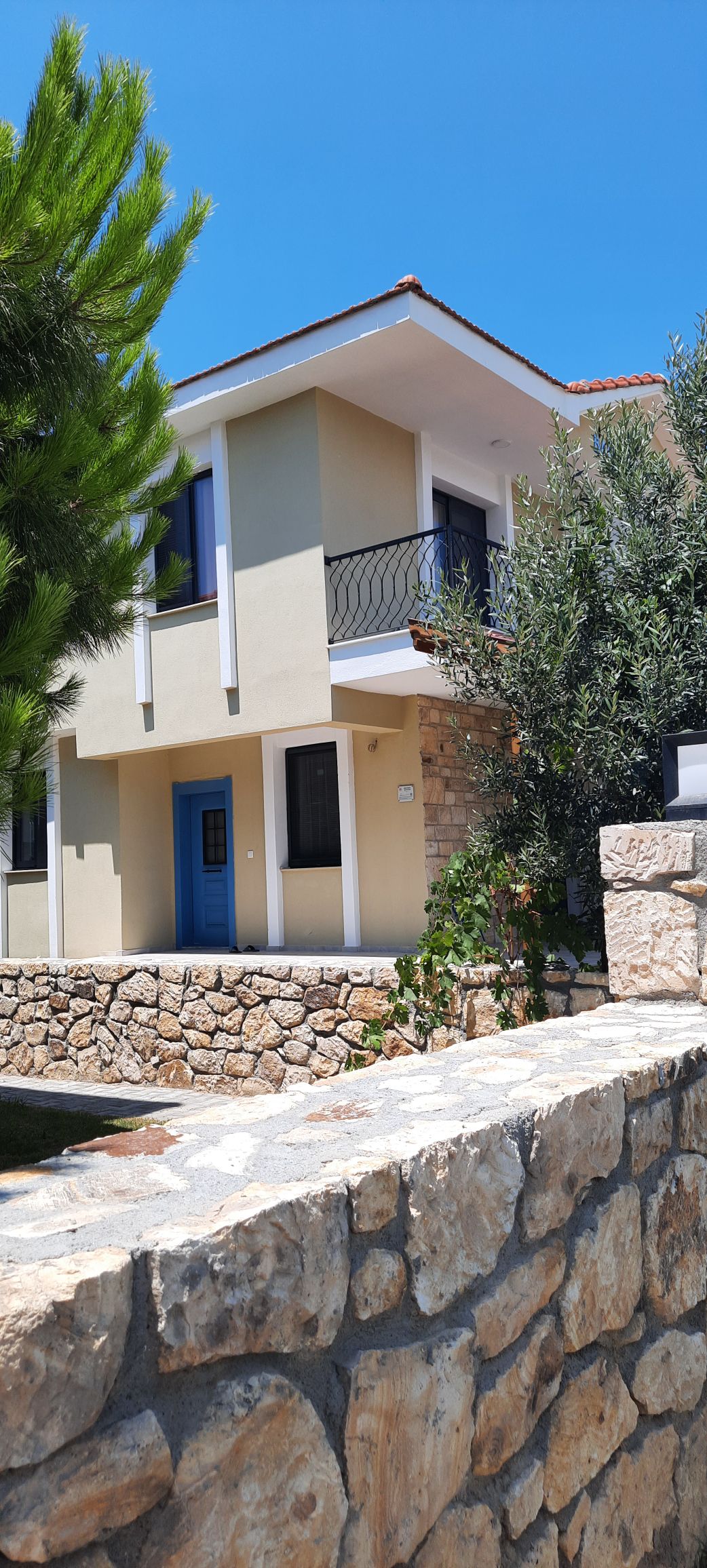 A two-story house with a stone facade and a vibrant blue door is surrounded by lush greenery. Large windows allow natural light to enter, while a small porch provides space for outdoor enjoyment. A stone wall borders the property, enhancing the overall landscaping.
