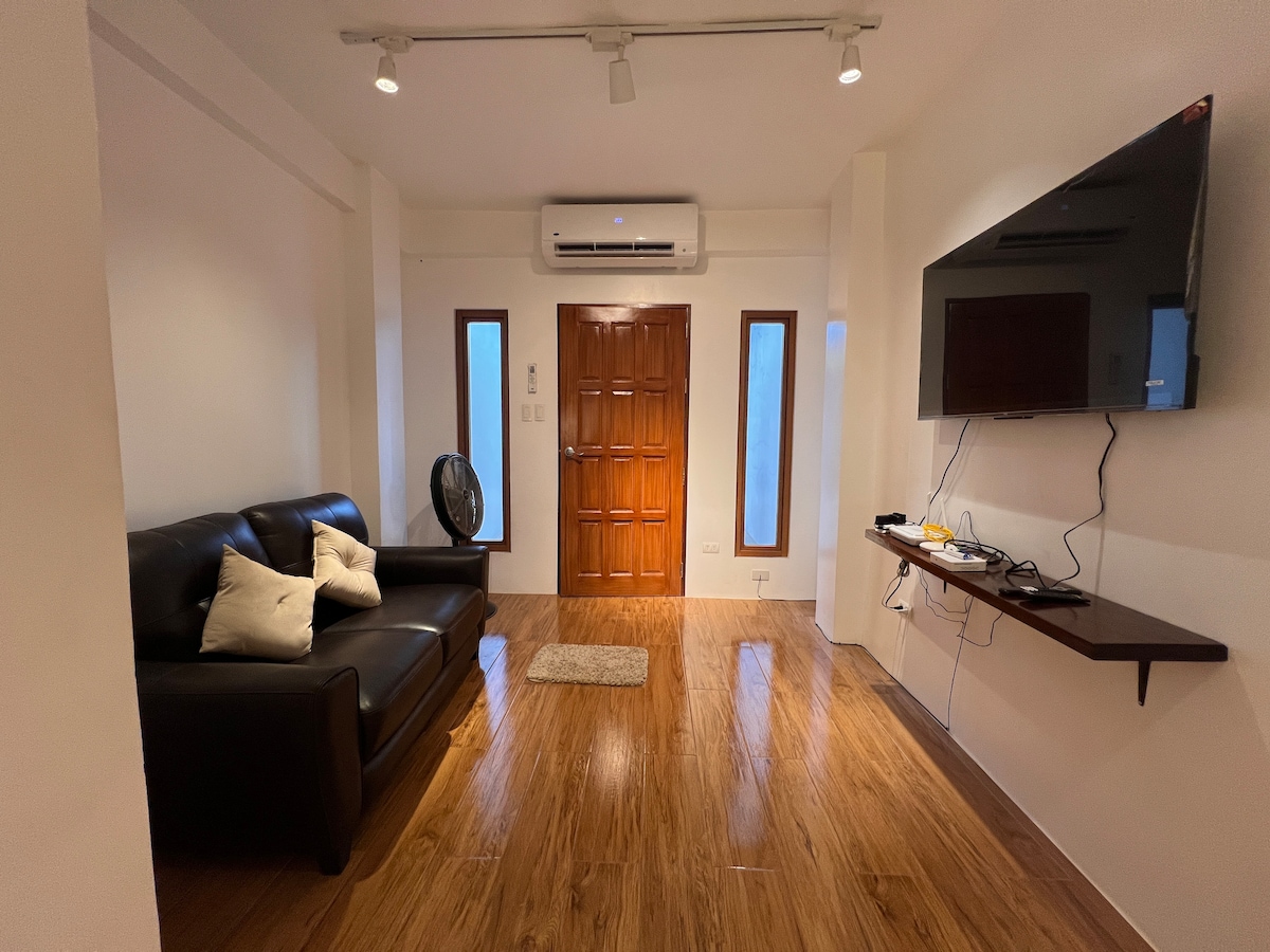 A well-lit living area features a black leather sofa adorned with two decorative pillows. A flat-screen television is mounted on the wall above a minimalist shelf. Warm wooden flooring complements the neutral tones of the walls, creating a comfortable space.