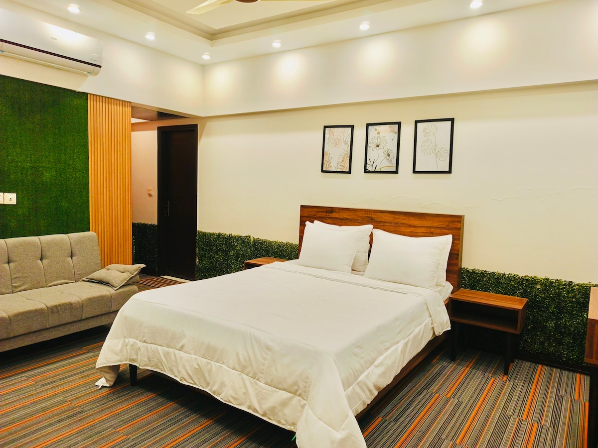 A spacious bedroom features a comfortable bed dressed in crisp white linens, complemented by wooden nightstands on either side. A cozy seating area with a gray sofa adds relaxation to the design. Green accents adorn the walls, enhancing the room's tranquil atmosphere.